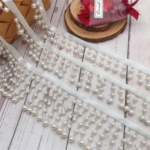 May include: White pearl trim with a satin ribbon backing. The trim features multiple rows of pearl beads hanging in a fringe style.