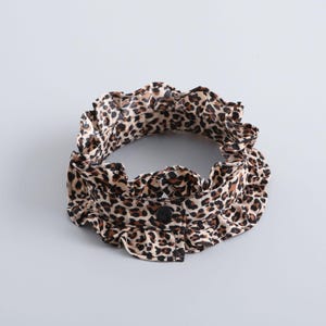 May include: A leopard print collar with a ruffled edge and a single black button. The collar is tan with brown spots and is set against a light gray background. The collar is a fashion accessory.