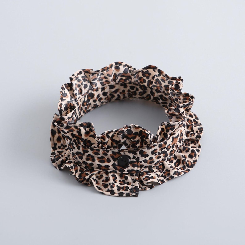 May include: A leopard print collar with a ruffled edge and a single black button. The collar is tan with brown spots and is set against a light gray background. The collar is a fashion accessory.