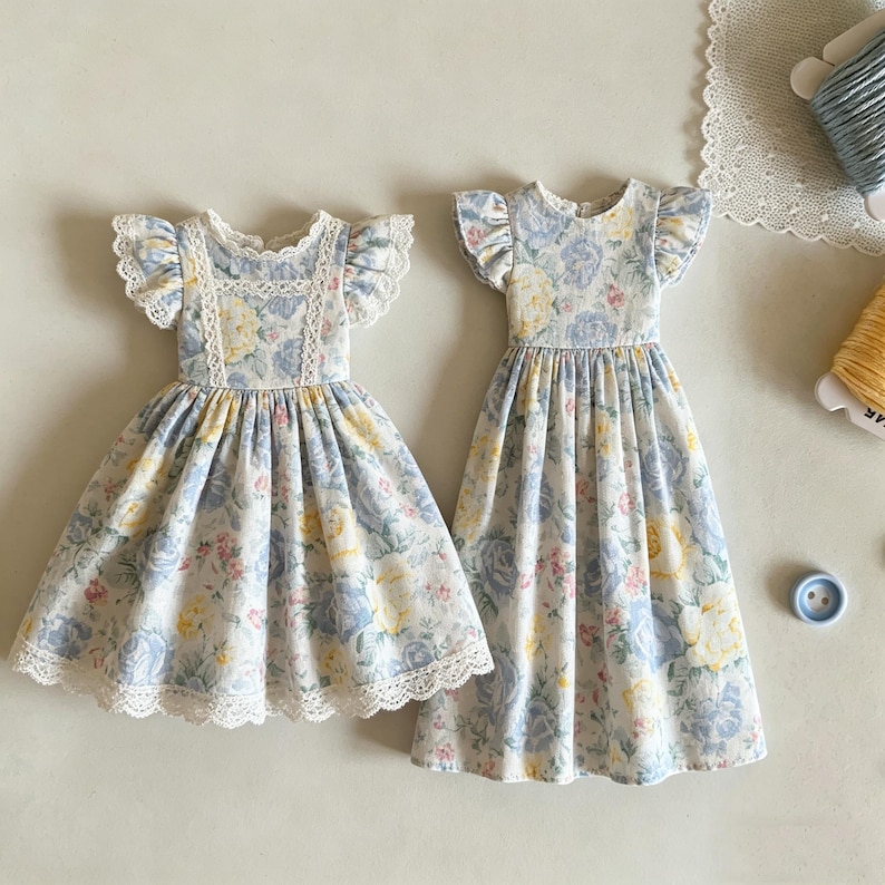 May include: Two floral print dresses with ruffled sleeves and lace trim. The dresses feature a light blue, yellow, and pink floral pattern on a white background. One dress has a shorter skirt, while the other is longer.