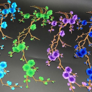 Può includere: Applicazioni ricamate di rami floreali nei toni del blu, verde e viola. I fiori sono dettagliati con più petali e i rami sono di colore marrone caldo. Le applicazioni sono su uno sfondo scuro.