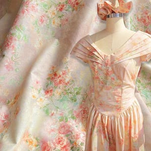 May include: A vintage-style dress and fabric featuring a floral pattern in soft pastel colors. The dress, displayed on a mannequin, has a pleated bodice and a full skirt. The fabric, with a similar rose design, drapes in the background, creating a cohesive aesthetic.