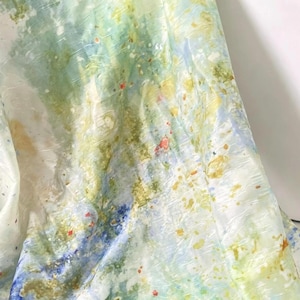 Pode incluir: Um tecido leve com uma estampa de aquarela abstrata em tons misturados de verde claro, azul suave e amarelo, acentuado por pequenas manchas laranja e azul escuro. O material apresenta uma textura sutilmente enrugada em toda a superfície.