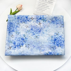 May include: A rectangular piece of fabric featuring a repeating pattern of blue hydrangea flowers and green leaves on a light blue background. The fabric has a soft, semi-transparent appearance with a delicate, layered floral design.