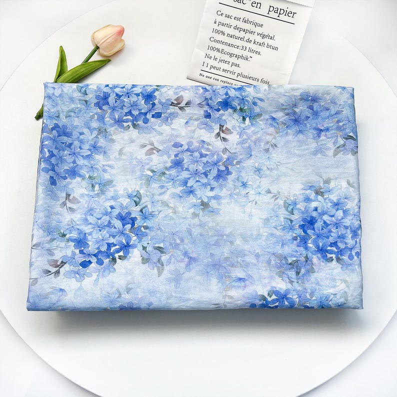 May include: A rectangular piece of fabric featuring a repeating pattern of blue hydrangea flowers and green leaves on a light blue background. The fabric has a soft, semi-transparent appearance with a delicate, layered floral design.