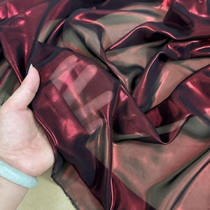 May include: A length of shimmering fabric in red and black hues. The material has a glossy, iridescent finish, with light reflecting off the folds. The fabric appears lightweight and flowing, with a translucent quality.