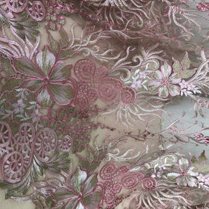 May include: Close-up of a delicate floral lace fabric. The design features intricate embroidery in shades of pink, white, and gold. The lace is semi-transparent, revealing the underlying fabric. The floral pattern includes flowers, leaves, and swirling details.