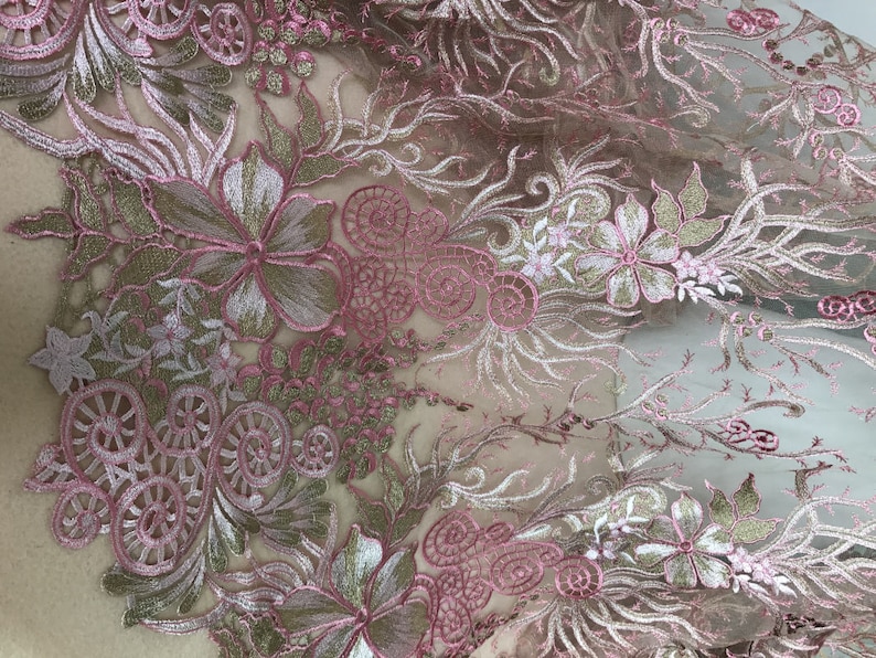 May include: Close-up of a delicate floral lace fabric. The design features intricate embroidery in shades of pink, white, and gold. The lace is semi-transparent, revealing the underlying fabric. The floral pattern includes flowers, leaves, and swirling details.