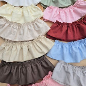 Op de afbeelding: Een collectie van meerdere off-shoulder gerimpelde stoffen banden in diverse effen kleuren, waaronder wit, crème, beige, bruin, lichtblauw, roze, rood en saliegroen. Elk stuk is voorzien van een geplooide elastische bovenrand en een uitlopende zoom.