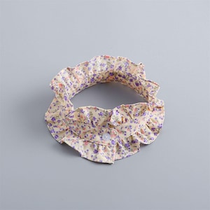 May include: A floral collar in a cream color with purple and pink flowers. The collar has a ruffled edge and a button closure. The collar is likely for a baby or child.