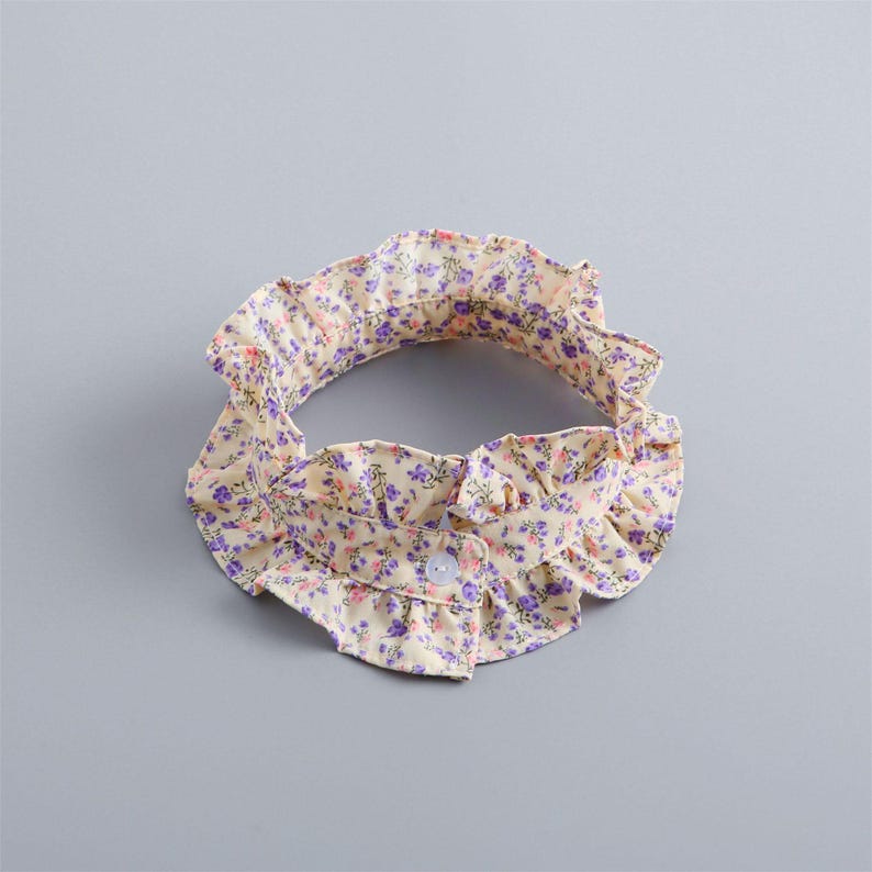 May include: A floral collar in a cream color with purple and pink flowers. The collar has a ruffled edge and a button closure. The collar is likely for a baby or child.