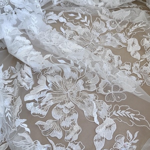 May include: White floral lace fabric with a delicate pattern of flowers and leaves. The lace is sheer and has a soft, flowing texture.