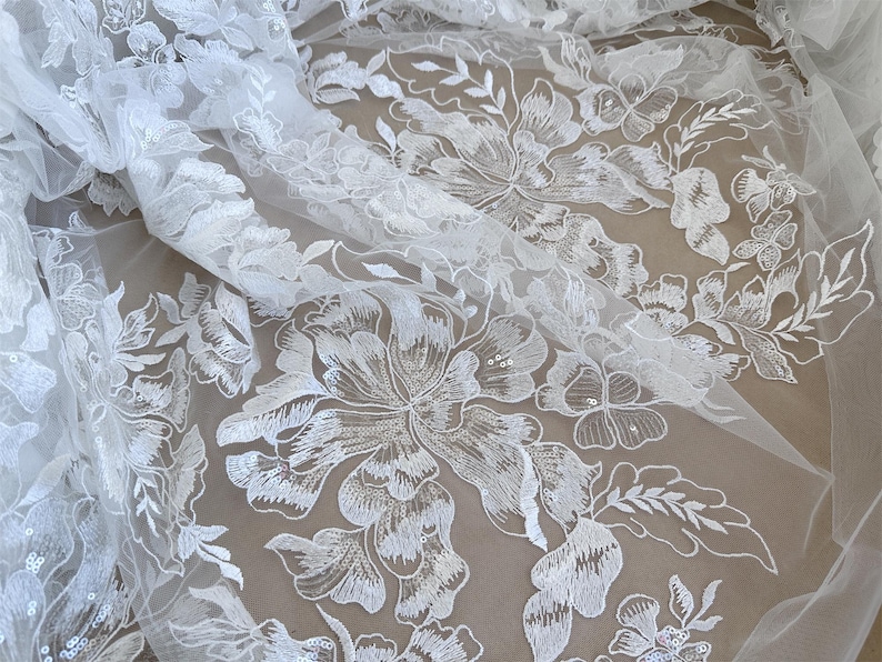 May include: White floral lace fabric with a delicate pattern of flowers and leaves. The lace is sheer and has a soft, flowing texture.
