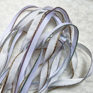 May include: Close-up of white ribbon with metallic gold, silver, and purple trim. The ribbon is arranged in a loose, overlapping pattern, creating a textured effect. The ribbon is likely used for crafting, sewing, or gift wrapping.