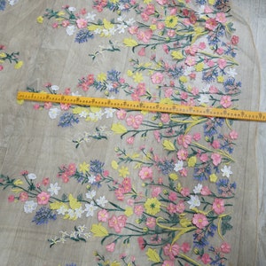 May include: A close-up of a sheer fabric with embroidered floral designs in shades of pink, yellow, white, and blue. The fabric is measured with a ruler.