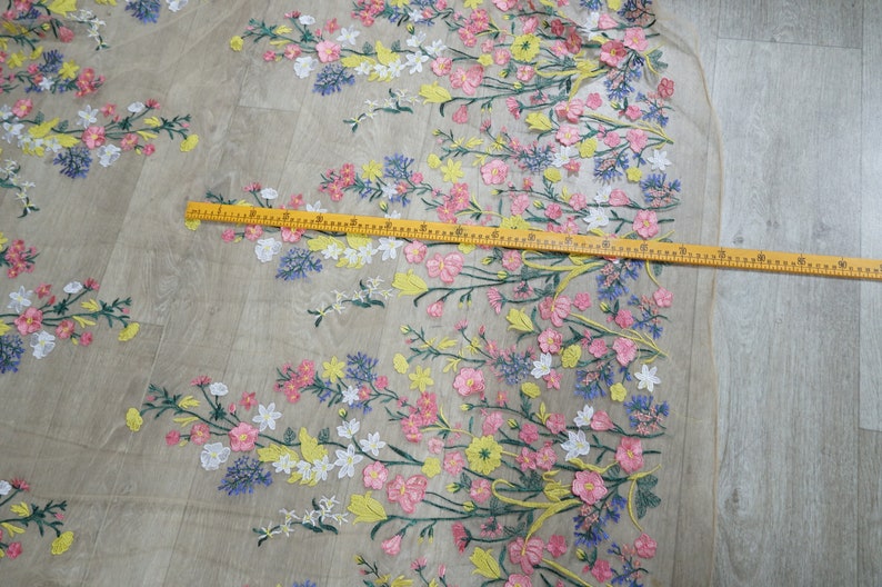 May include: A close-up of a sheer fabric with embroidered floral designs in shades of pink, yellow, white, and blue. The fabric is measured with a ruler.