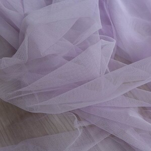 May include: A close-up of a soft, light purple tulle fabric. The fabric is delicate and sheer, with a fine mesh weave.