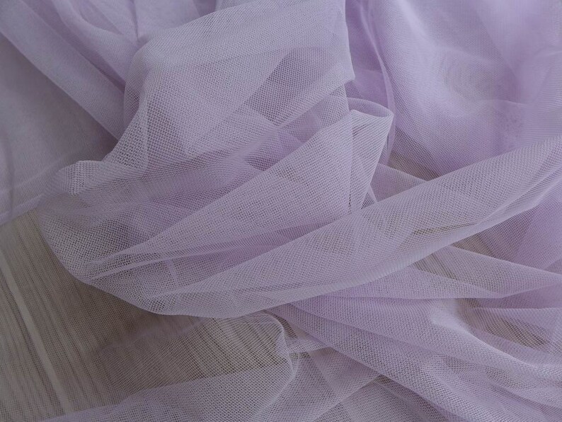 May include: A close-up of a soft, light purple tulle fabric. The fabric is delicate and sheer, with a fine mesh weave.