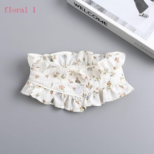 May include: A white floral collar with a ruffled edge and a small bow. The collar features a delicate pattern of small flowers in shades of pink, blue, and green. It fastens with a single button.