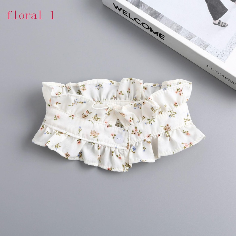 May include: A white floral collar with a ruffled edge and a small bow. The collar features a delicate pattern of small flowers in shades of pink, blue, and green. It fastens with a single button.