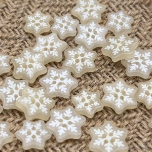以下が含まれることがあります： 6つの突起を持つ雪の結晶の形をした、半透明のオフホワイトのボタンのコレクション。各ボタンの表面には白い雪の結晶の模様が浮き彫りになっており、中央には縫い付け用の4つの小さな穴が開いています。