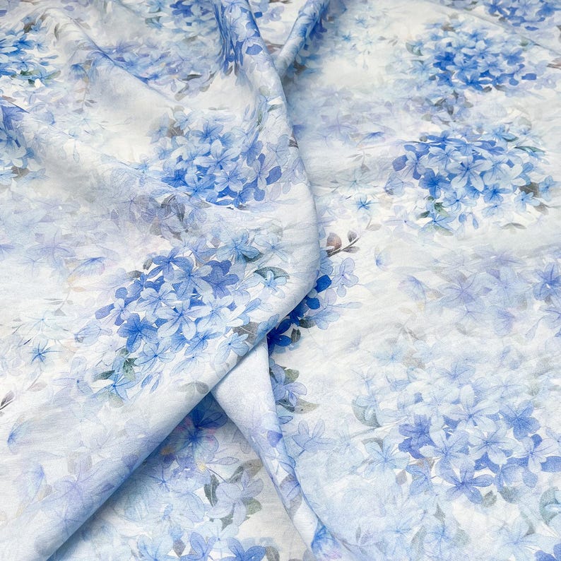 May include: A lightweight fabric featuring a repeating watercolour-style print of blue hydrangea clusters on a white background. The material is draped to show folds and texture, highlighting the soft blue and white floral pattern throughout.