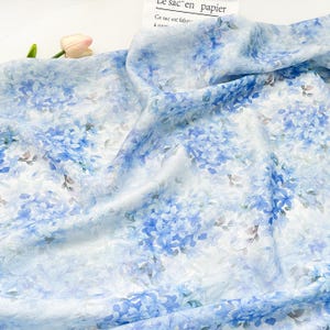 May include: A lightweight, semi-sheer fabric featuring a repeating pattern of blue hydrangea clusters on a white background. The material is draped in soft folds, showcasing the delicate floral print and the subtle variation in blue tones.
