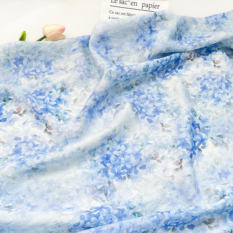 May include: A lightweight, semi-sheer fabric featuring a repeating pattern of blue hydrangea clusters on a white background. The material is draped in soft folds, showcasing the delicate floral print and the subtle variation in blue tones.