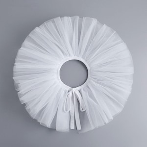 Può includere: Una gonna tutù bianca realizzata con strati di tessuto di tulle trasparente. La gonna ha una forma circolare con una grande apertura al centro e una chiusura con fiocco. Il tulle è raccolto per creare un aspetto pieno e soffice.