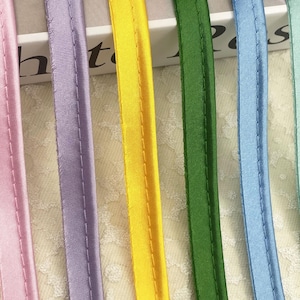 May include: Six vertical strips of satin piping in pink, lavender, yellow, dark green, light blue, and mint green. Each strip features a central raised cord encased in fabric with a single line of stitching running parallel along the side.
