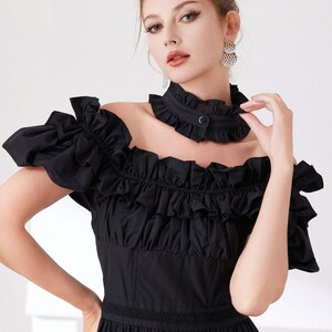 May include: Black off-the-shoulder dress with ruffled sleeves and bodice. The dress features a high, ruffled collar with a button closure. The skirt is tiered, and the dress has a fitted waist. The model is wearing silver earrings.