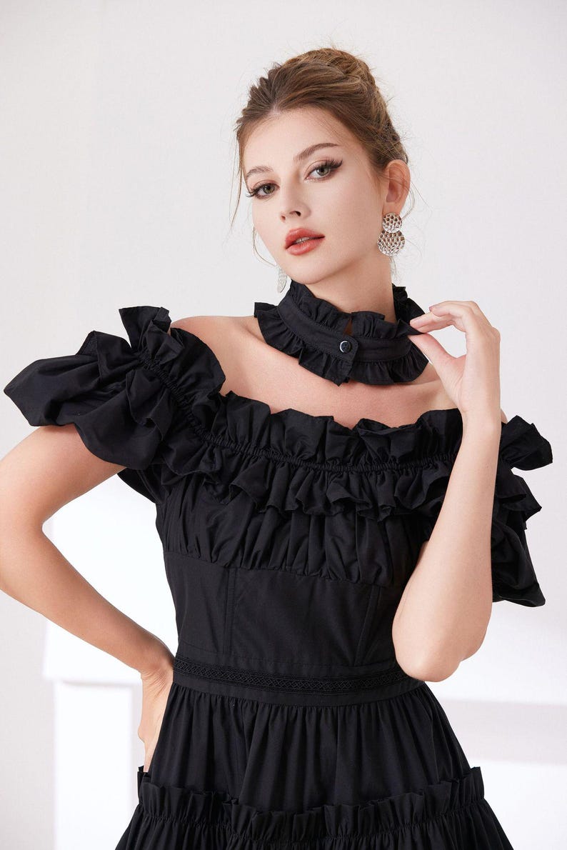 May include: Black off-the-shoulder dress with ruffled sleeves and bodice. The dress features a high, ruffled collar with a button closure. The skirt is tiered, and the dress has a fitted waist. The model is wearing silver earrings.