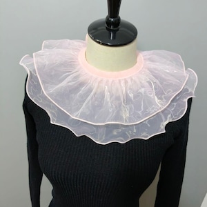 May include: A detachable collar featuring two layers of sheer, pale pink organza fabric with a ruffled, wavy edge. The collar is constructed with a solid, light pink fabric band at the neckline and is displayed on a black mannequin torso.