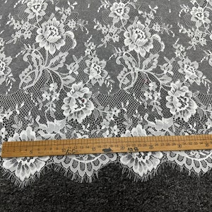 May include: White floral lace fabric with a delicate pattern. The lace has a scalloped edge and is measured with a ruler in inches.