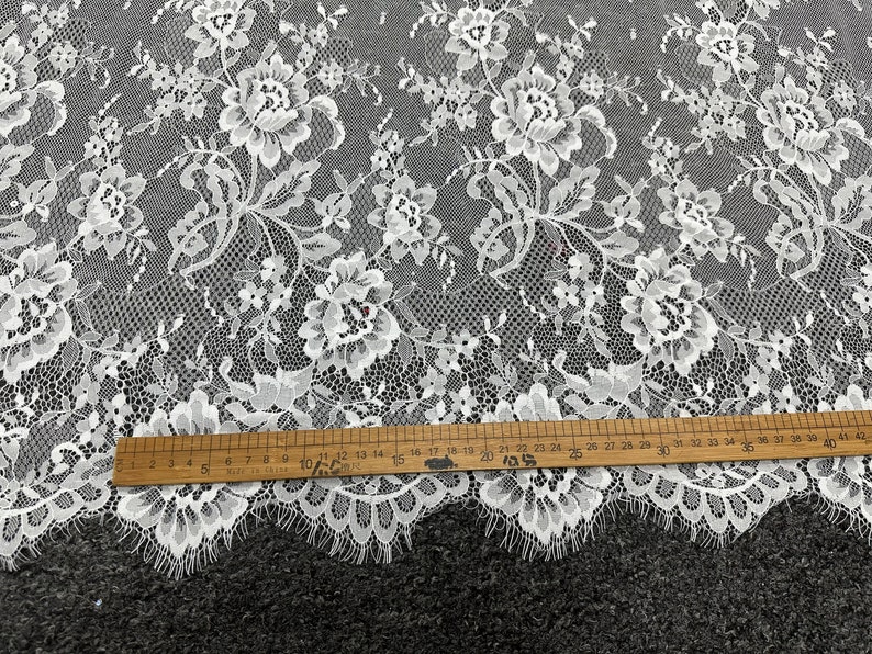 May include: White floral lace fabric with a delicate pattern. The lace has a scalloped edge and is measured with a ruler in inches.