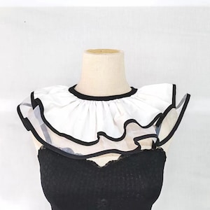 May include: A white detachable collar featuring two layers of ruffled fabric with black trim along the edges. The collar is displayed on a mannequin neck and shoulders, showcasing a rounded neckline and a layered, pleated design with black piping.