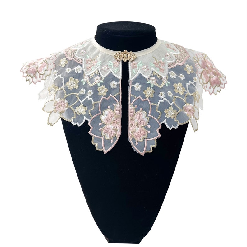 May include: A white lace collar with pink floral embroidery. The collar has a scalloped edge and a delicate design.