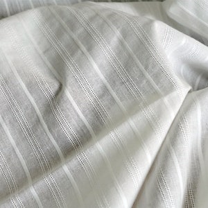 May include: White fabric with thin, vertical stripes. The fabric is woven with a subtle texture and has a slightly sheer appearance.