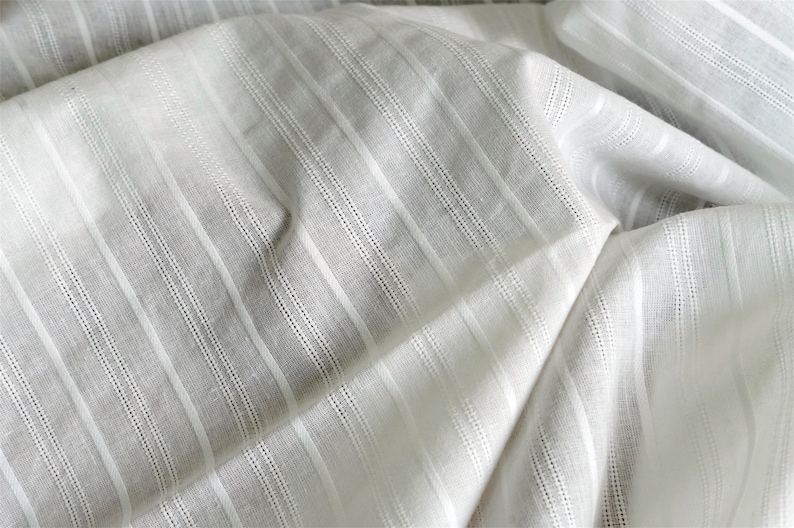 May include: White fabric with thin, vertical stripes. The fabric is woven with a subtle texture and has a slightly sheer appearance.