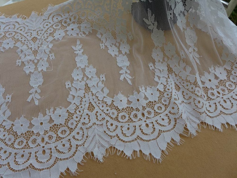 Puede incluir: Borde de encaje blanco con un delicado patr&oacute;n floral y un borde festoneado. El encaje es transparente y tiene una textura suave y fluida.