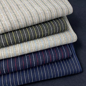 Op de afbeelding: Een stapel van vijf gevouwen stofstalen met krijtstreepmotieven. De kleuren omvatten lichtgrijs met witte strepen, donker antraciet met gele strepen, lichtblauw met witte strepen, marineblauw met rode strepen en marineblauw met witte strepen.