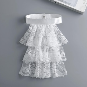 May include: A white lace collar with three tiered ruffles and a satin band. The collar is fastened with a single button and is set against a gray background. A book is visible in the background.