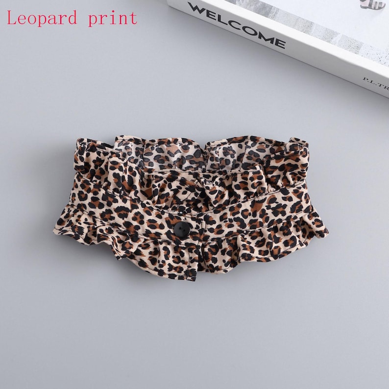 May include: A leopard print accessory with a ruffled edge and a single black button closure. The fabric features a pattern of brown and black spots on a beige background. The text "leopard print" is visible in the image.