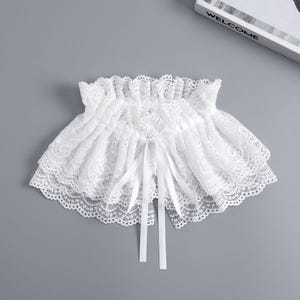 May include: A white lace collar with a ruffled design and a satin ribbon tie. The collar is made of delicate lace with a scalloped edge. The word "WELCOME" is visible on a white book in the background.