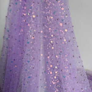 May include: A sheer lavender fabric adorned with iridescent sequins and small, round beads. The fabric is draped, creating soft folds and shadows. The sequins catch the light, adding a shimmering effect. This material is suitable for creating elegant apparel.