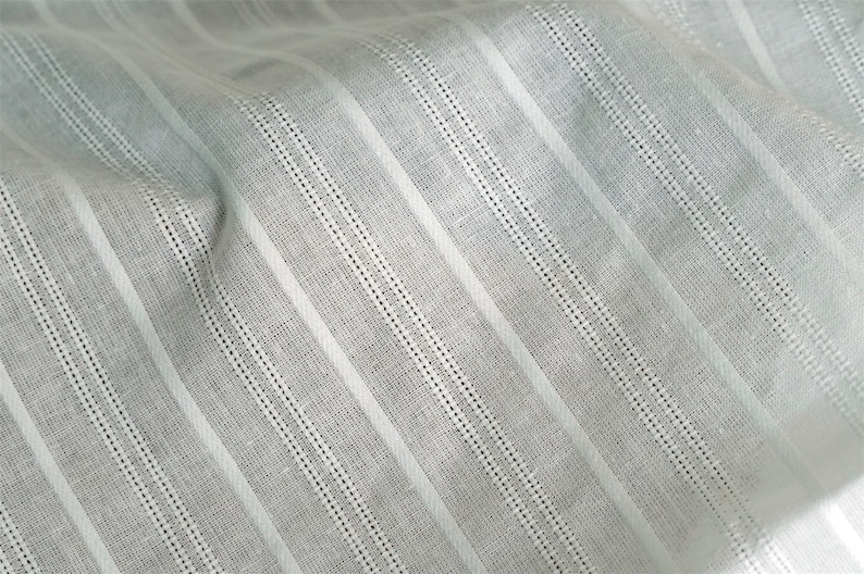 May include: White fabric with thin, vertical stripes. The fabric has a subtle texture and a slightly wrinkled appearance.