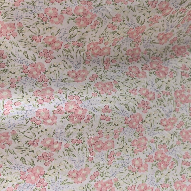 Puede incluir: Primer plano de una tela con estampado floral. El patr&oacute;n presenta grupos de flores rosas con hojas verdes y peque&ntilde;as flores azules sobre un fondo blanco. La tela tiene una est&eacute;tica delicada y vintage.