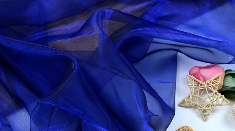 May include: A close-up of a royal blue sheer fabric with a soft, flowing texture. The fabric is draped over a white surface and has a subtle shimmer.