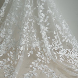 May include: White floral lace fabric with a delicate pattern of leaves and flowers. The lace is sheer and has a soft, flowing texture.