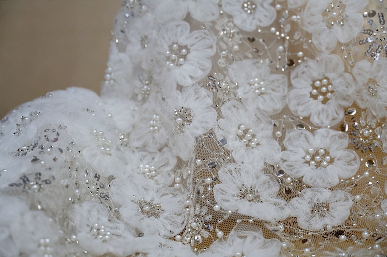 以下が含まれることがあります： 繊細な花柄のアップリケとパールがあしらわれた白いレース生地。生地は透き通っており、柔らかく、流れるような質感です。